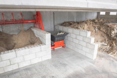 Ein orangefarbener Shredder in mitten von großen Betonbausteinen mit Förderschnecke zwischen einem Stapel Restholz und einem Bunker mit Hackschnitzeln