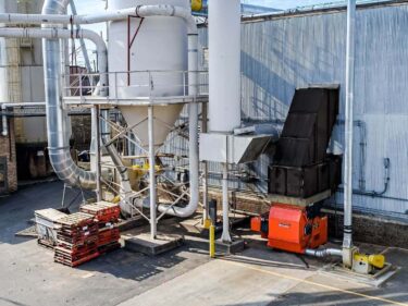 An orange WEIMA shredder is positioned behind a warehouse, surrounded by an air system with a white silo and metal duct work.