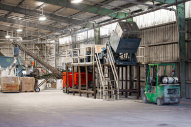 Eine industrielle Shredderanlage mit Zuführsystem und Förderband ist in einer geräumigen Werkhalle aufgebaut.
