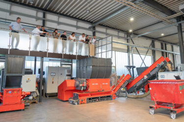 a WEIMA shredder in a large hall, people standing on a balcony looking at the machine